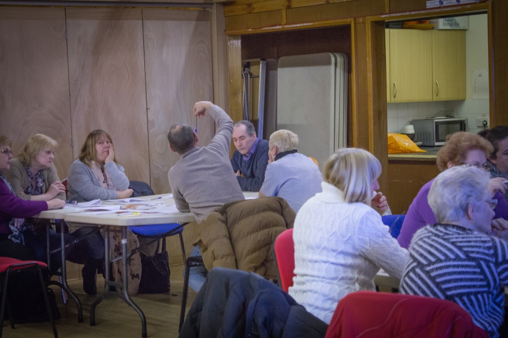 This photograph is of a GoWell Principle Investigator with the GoWell Panel - eight of the Panel are in the photograph. Five Panel members are sat at a table with the Principle Investigator reviewing his evidence. Three Panel members in the foreground of the picture are sat at a table preparing a role play exercise.  They are all at a community venue in Drumchapel, Glasgow.